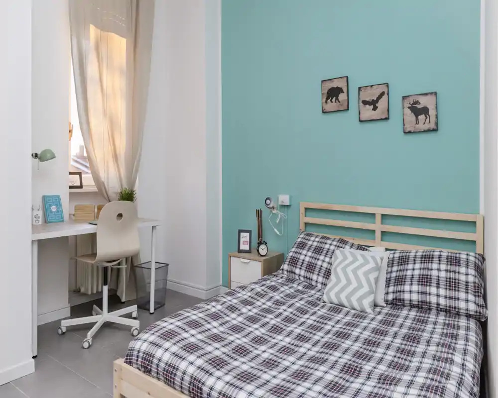 Bright, modern bedroom with a wooden bedframe, patterned duvet and a small workspace by the window. Calm teal accent wall and simple decorative art create a cozy, well-styled atmosphere ideal for short-term rentals.