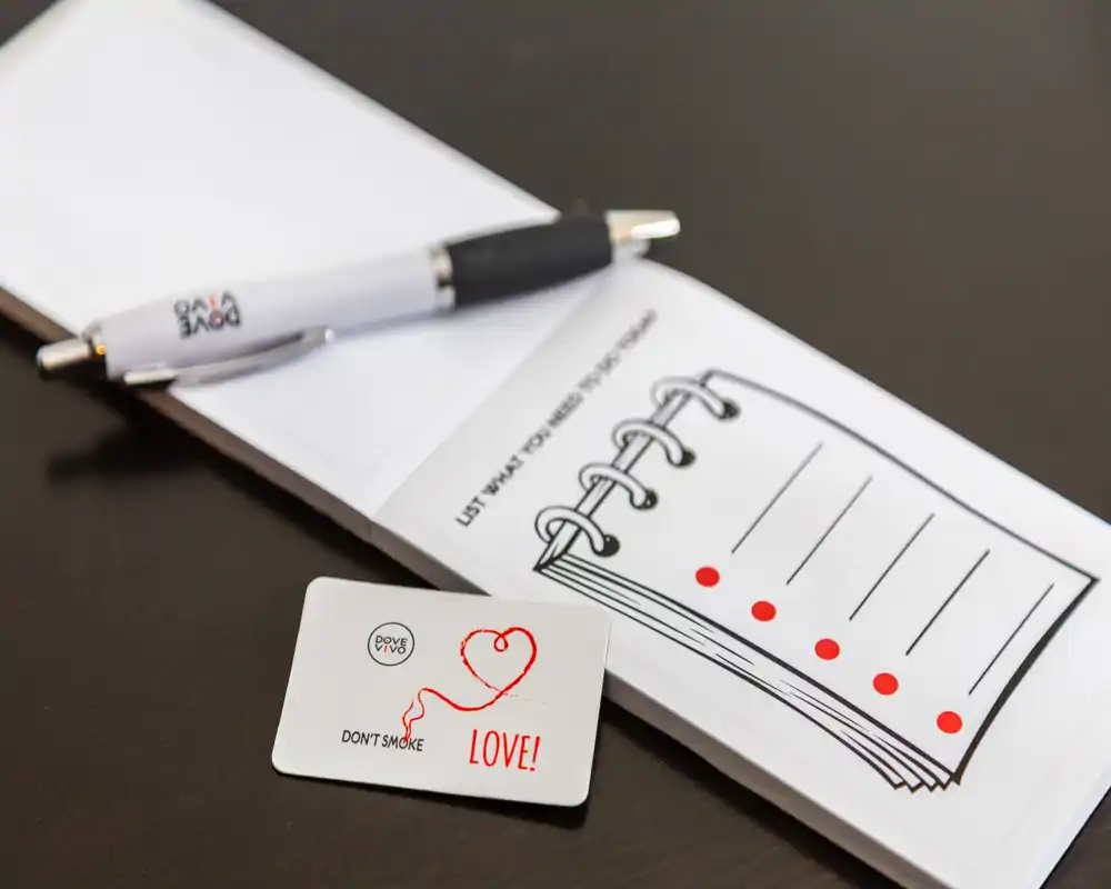 Primer plano de un bolígrafo, un bloc de notas pequeño y una tarjeta decorativa sobre una superficie oscura. La imagen destaca material de papelería y puede usarse para mostrar detalles de bienvenida o accesorios de escritorio.