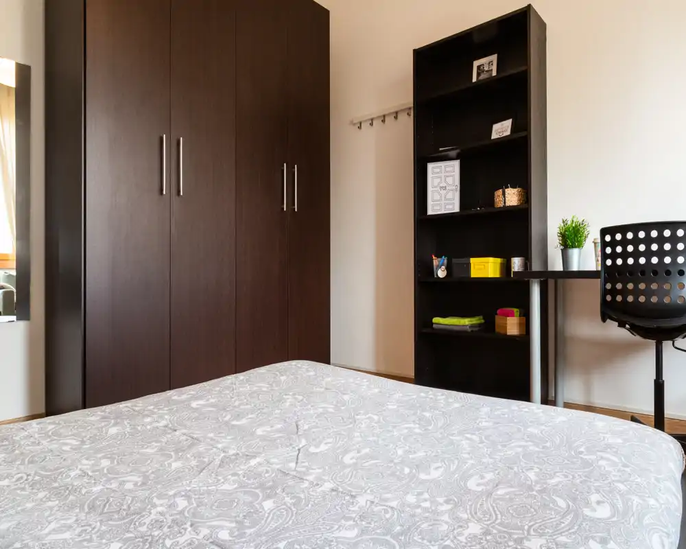 Dormitorio luminoso y de estilo sencillo con una gran cama en primer plano, armario y estantería de madera oscura y un pequeño escritorio junto a la ventana—limpio y funcional para estancias cortas.