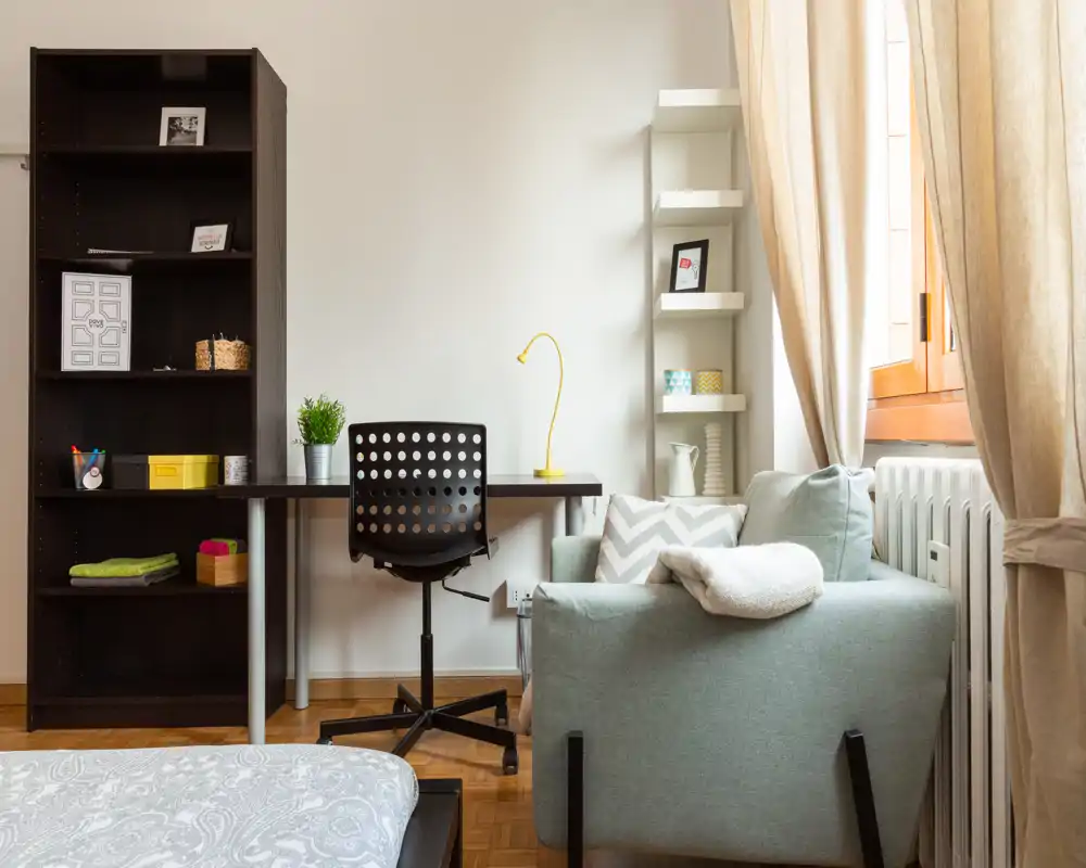 Rincón de dormitorio luminoso y bien decorado con borde de la cama visible, escritorio y un sillón cómodo junto a la ventana que crea un espacio acogedor con luz natural.