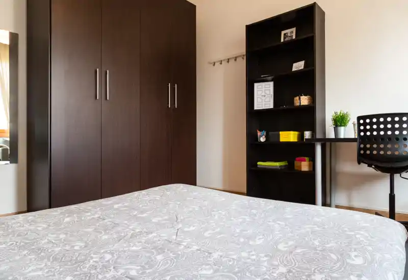 Bright, simply styled bedroom with a large bed in the foreground, dark wood wardrobe and shelving, and a small desk by the window—clean and functional for short stays.