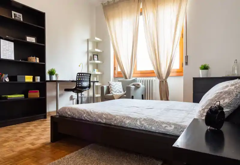 Bright, modern bedroom with a large bed in the foreground, a cozy armchair by the window, a desk and shelving for storage — ideal for comfortable stays.