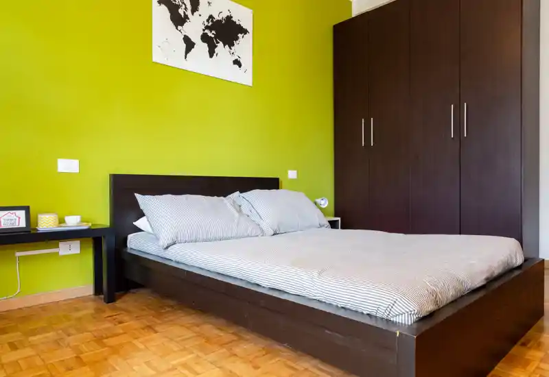 Bright, modern bedroom with a low dark-wood platform bed, striped bedding and a large wardrobe. The green accent wall and parquet floor give the room a fresh, contemporary feel.