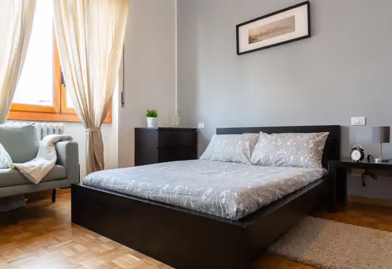 Bright, well-styled bedroom with a double bed, bedside table, chest of drawers and a cozy armchair by the window; neutral grey tones and natural light create a calm atmosphere.