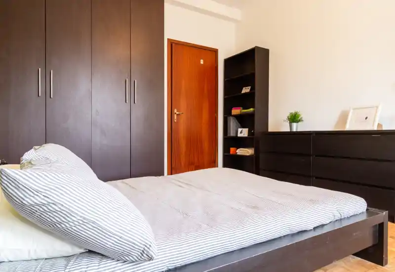 Bright, modern bedroom with a double bed in the foreground, dark wood wardrobes and matching dresser, and simple decorative accents creating a clean, comfortable space.