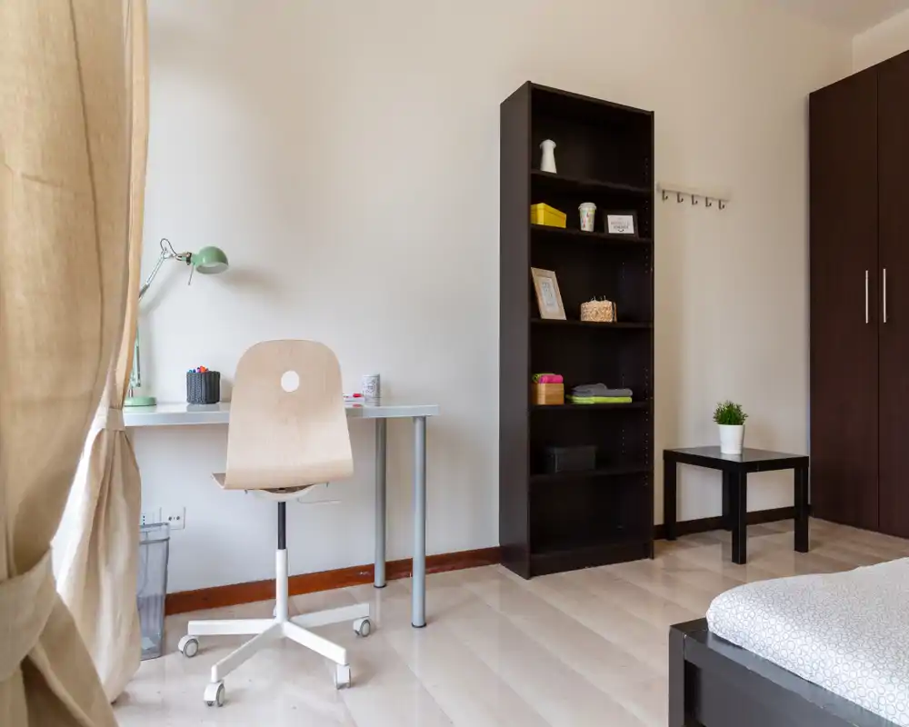Angolo camera luminoso e minimal con scrivania e sedia, una libreria scura e il letto parzialmente visibile in primo piano. La luce naturale dalla finestra crea un'atmosfera tranquilla e ariosa, perfetta per un annuncio di affitto.
