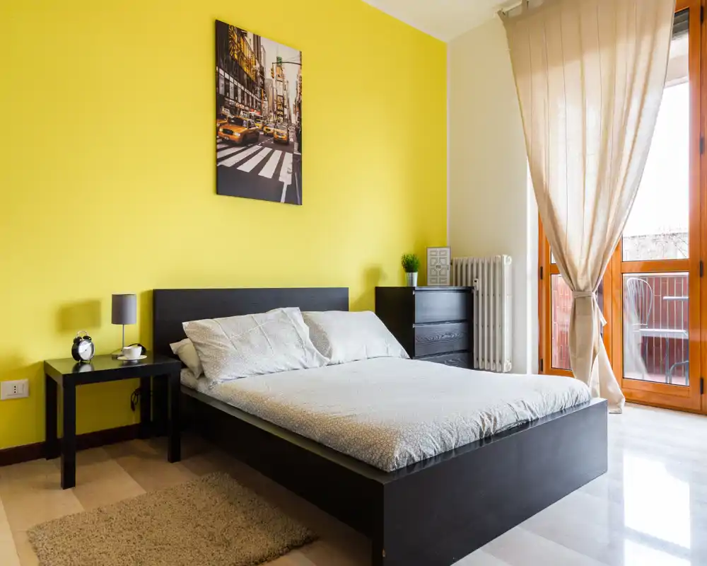 Camera da letto moderna e luminosa con letto matrimoniale, comodini, decor minimal e porta sul balcone che illumina la stanza — accogliente e ordinata per affitti brevi.