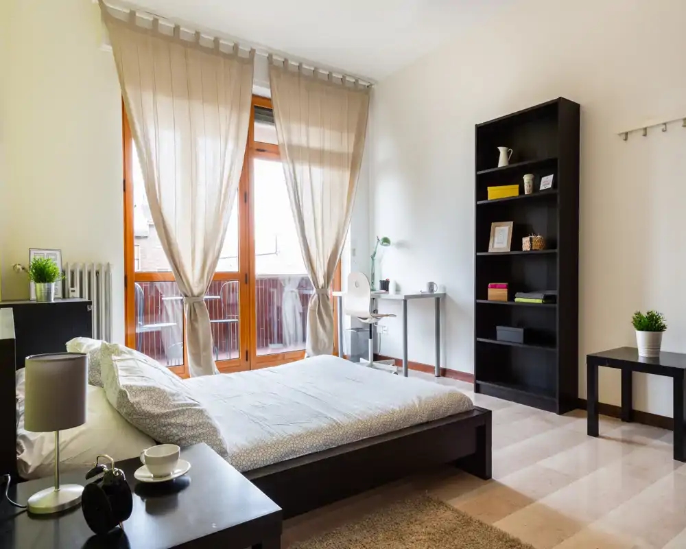 Camera da letto luminosa e ben arredata con letto matrimoniale di fronte alle ampie porte del balcone. Finestre con cornice in legno e tende chiare illuminano l’ambiente; scaffale e una piccola scrivania completano la disposizione moderna e funzionale.