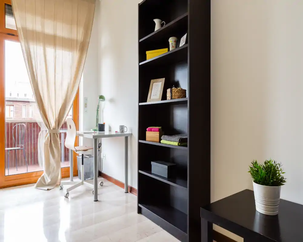 Angolo della camera luminoso e minimal con una piccola scrivania, libreria e porta sul balcone che lascia entrare luce naturale; stile moderno e ordinato, adatto a una persona sola.