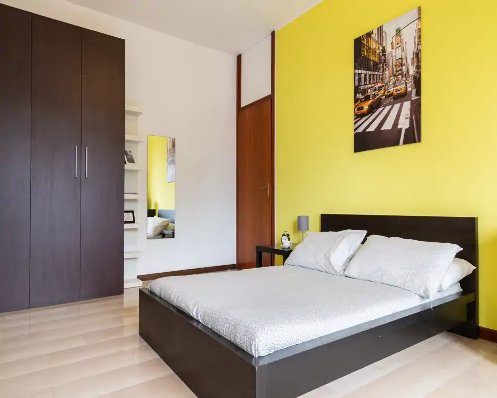 Camera da letto luminosa e spaziosa con letto basso matrimoniale, biancheria pulita e parete gialla a contrasto. Ampio armadio e comodino minimal completano l’arredamento moderno e ordinato, perfetto per annunci.