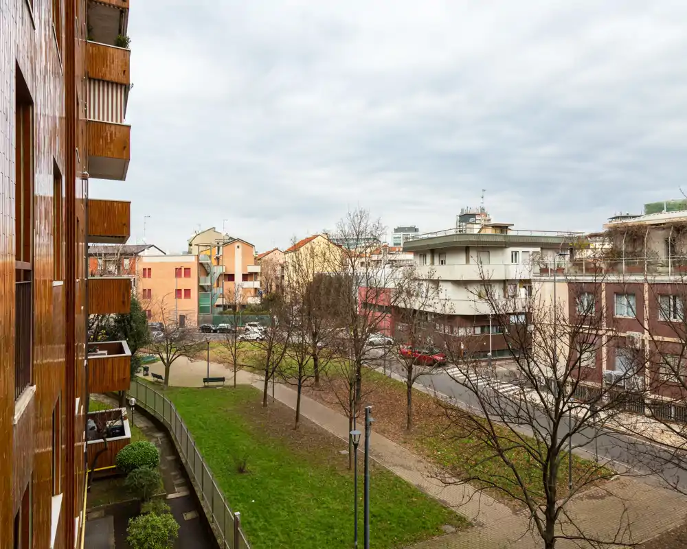 Vista desde el balcón hacia un patio residencial y la calle con árboles, césped y edificios vecinos bajo un cielo nublado.