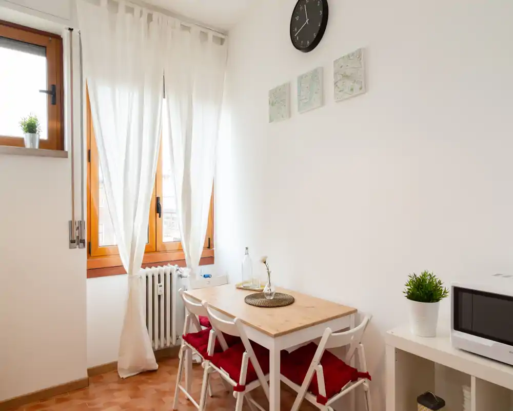 Acogedor rincón de comedor luminoso con mesa de madera, cuatro sillas plegables blancas con cojines rojos, luz natural de una gran ventana y detalles decorativos sencillos.