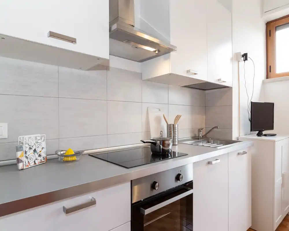 Cocina moderna y compacta con placa de inducción, horno empotrado y fregadero de acero inoxidable. Superficies limpias y bien iluminadas con almacenamiento práctico.