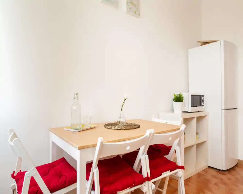Zona de comedor compacta y luminosa con mesa de madera, cuatro sillas plegables blancas con cojines rojos, un bajo plato decorativo y un pequeño jarrón; frigorífico y microondas visibles junto a la mesa.