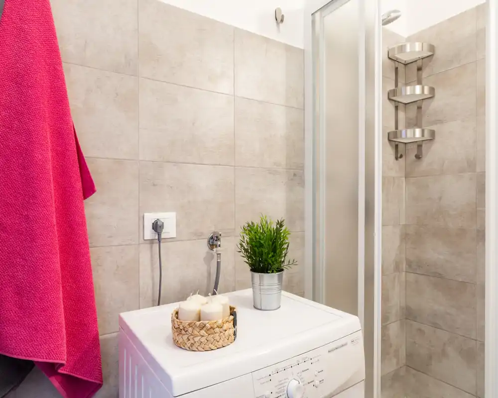 Baño moderno y compacto con paredes alicatadas, cabina de ducha y lavadora de carga superior con velas decorativas y una planta pequeña encima.