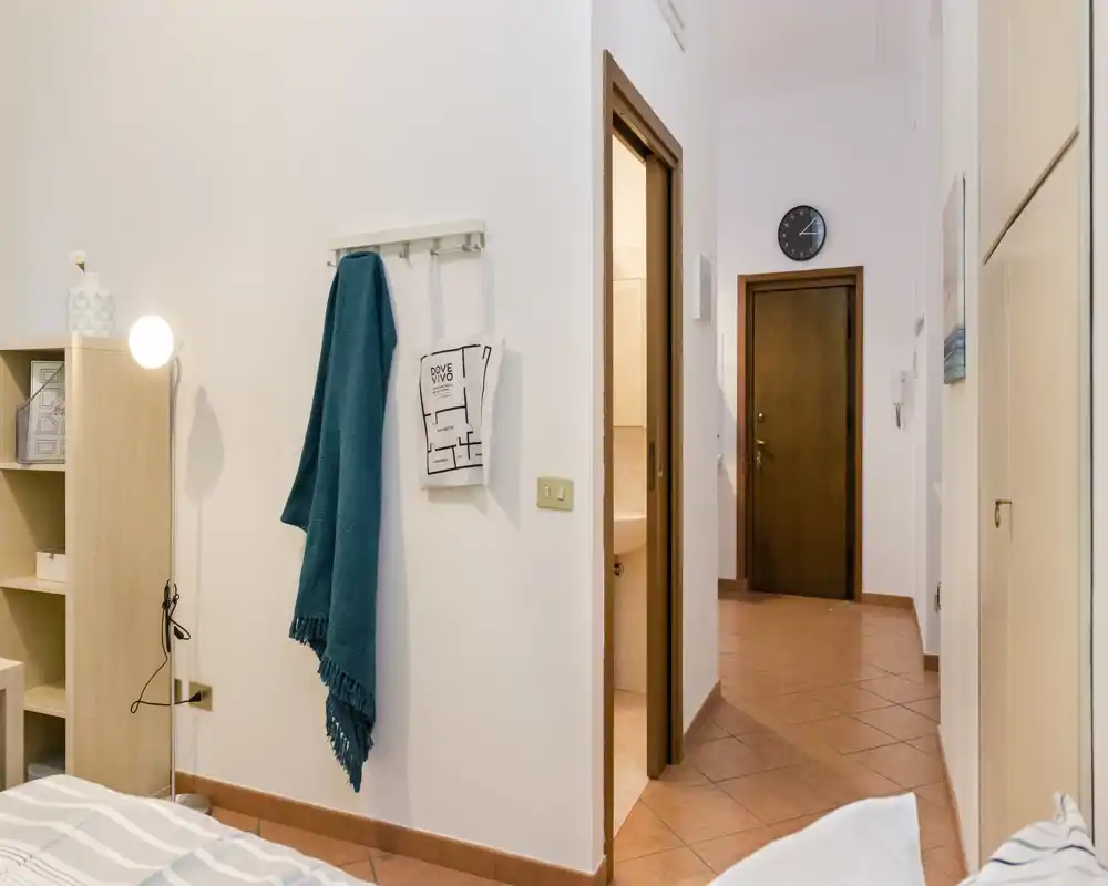 Rincón de dormitorio luminoso y ordenado con ropa de cama visible, estantería junto a la cama y una lámpara de pie; al fondo se aprecia el pasillo y la puerta de entrada.