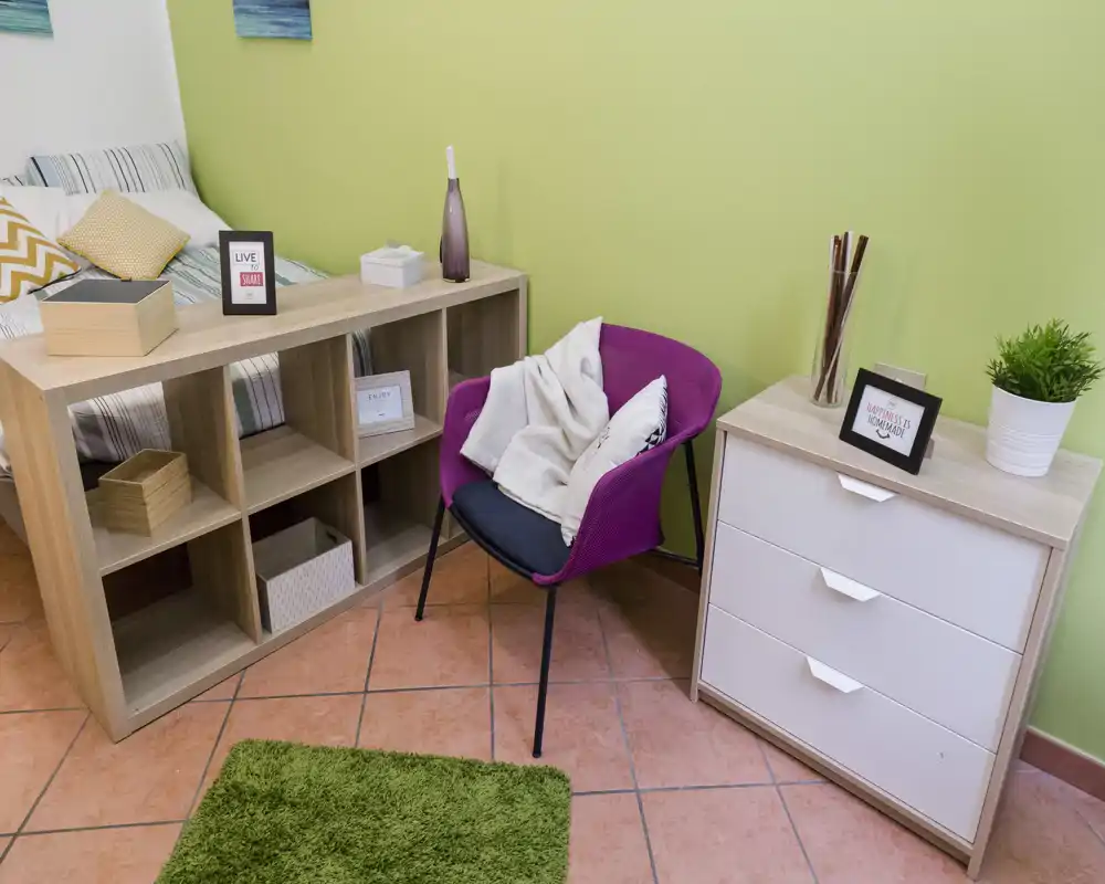Rincón de dormitorio luminoso y acogedor con cama visible, estantería, sillón y cómoda — ideal para anuncios de estudios o apartamentos pequeños.