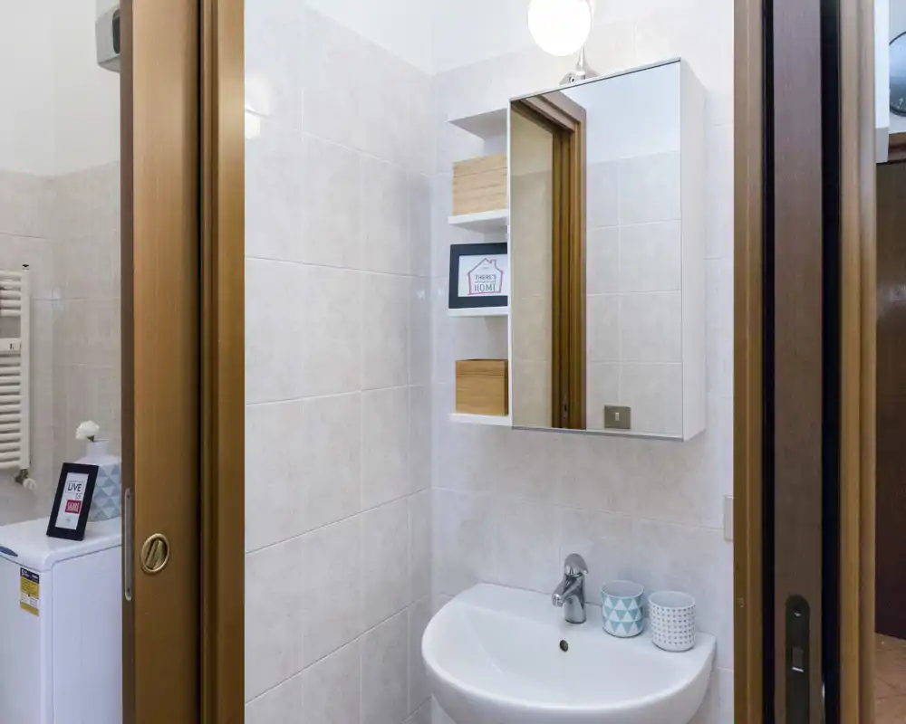 Pequeño baño alicatado con mueble espejo, lavabo blanco y grifo moderno; tazas decorativas en el lavabo y estantes que ofrecen un aspecto ordenado y bien iluminado.