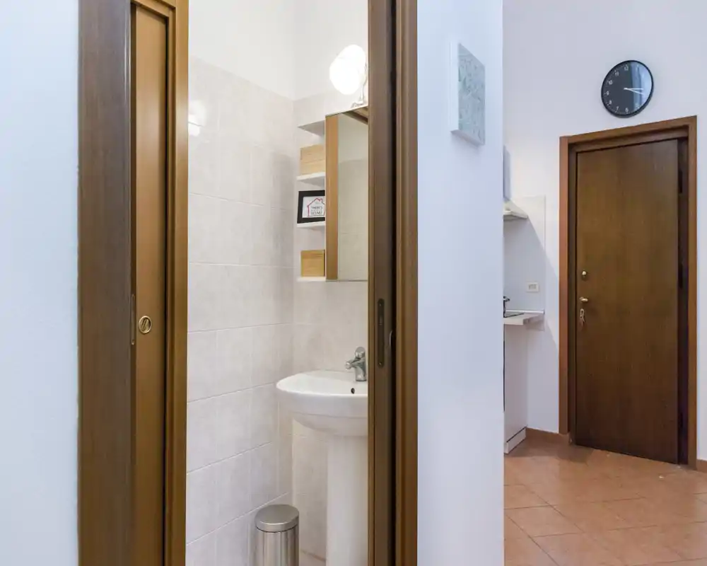 Baño compacto con lavabo de pedestal, armario con espejo y azulejos claros; visible a través de una puerta corredera de madera junto a la entrada.