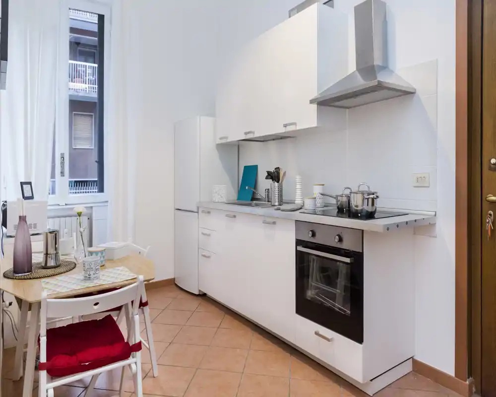 Cocina moderna y compacta con muebles blancos, horno empotrado y una pequeña mesa junto a una ventana luminosa — ideal para un piso urbano.