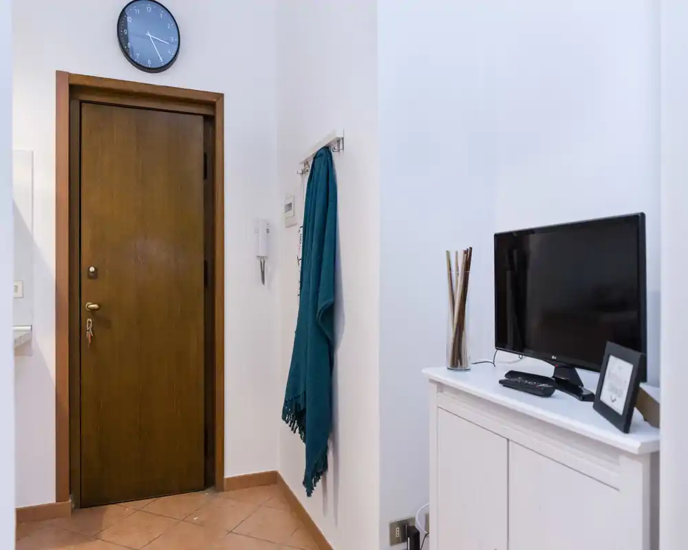 Pequeño y ordenado recibidor con puerta de madera, perchas con un chal colgado, reloj de pared y una consola con televisor y jarrón decorativo.
