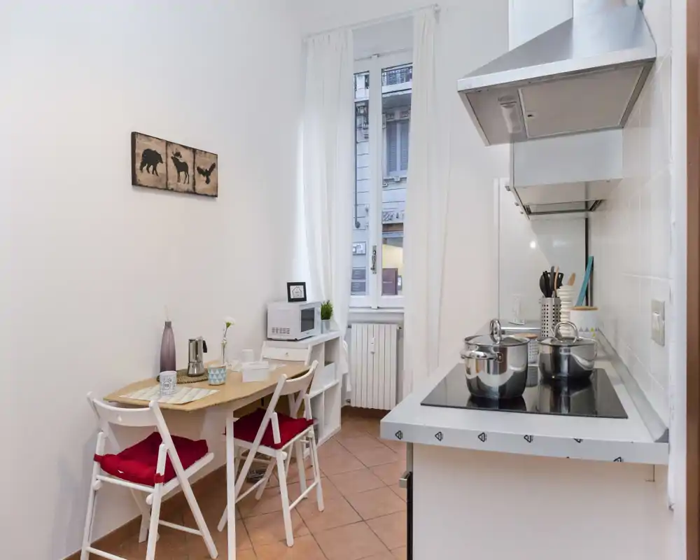 Cocina compacta y luminosa con placa moderna, ollas en la vitrocerámica y una pequeña mesa con sillas con cojines rojos—perfecta para comidas acogedoras.
