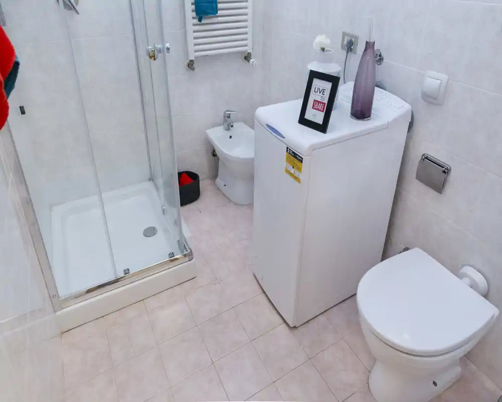 Baño compacto y limpio con cabina de ducha, inodoro, bidé y lavadora; azulejos en paredes y suelo que dan un espacio luminoso y funcional.