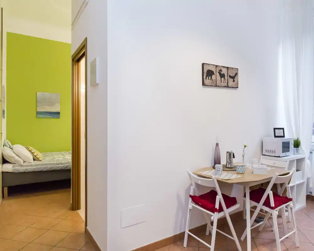 Vista de un estudio: dormitorio visible a través de una puerta abierta y un pequeño rincón comedor con mesa, sillas y microondas. El dormitorio tiene una cama con cojines y una pared verde de acento.