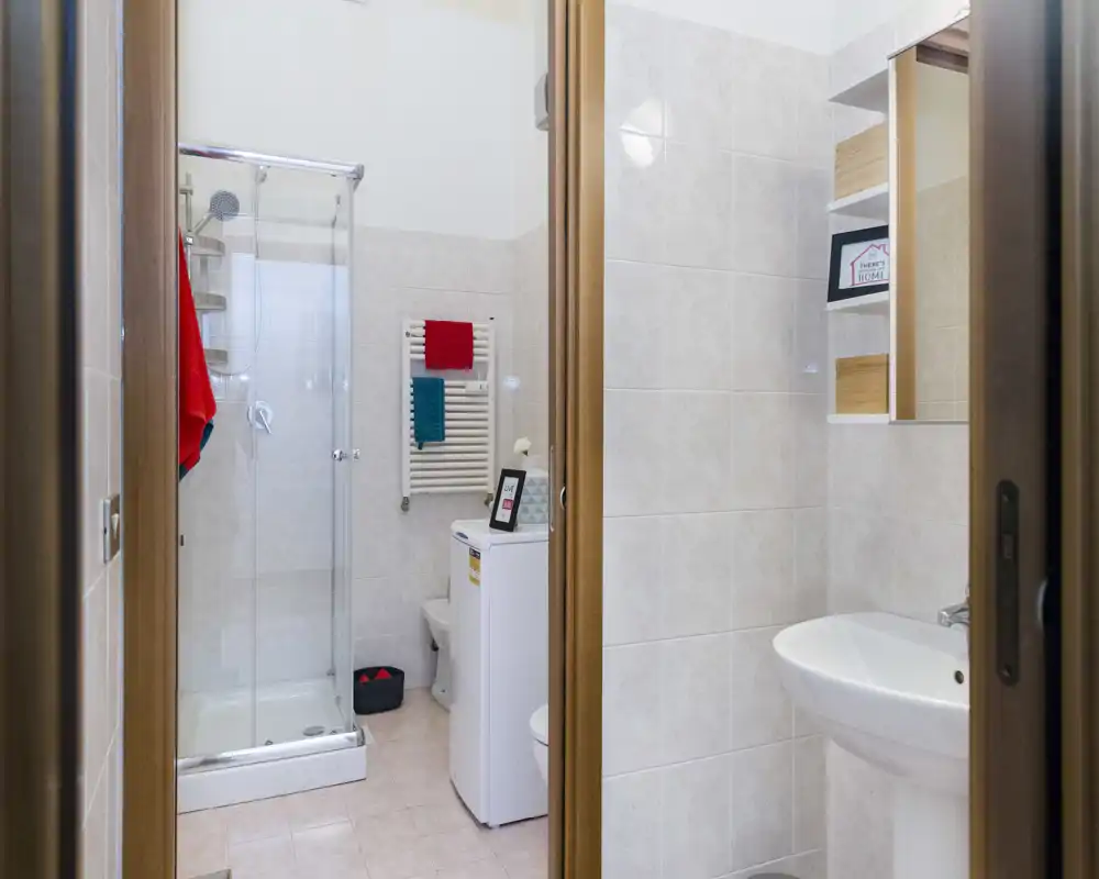 Baño moderno y luminoso con ducha de cristal, radiador toallero, lavabo y lavadora; paredes alicatadas claras y decoración ordenada.