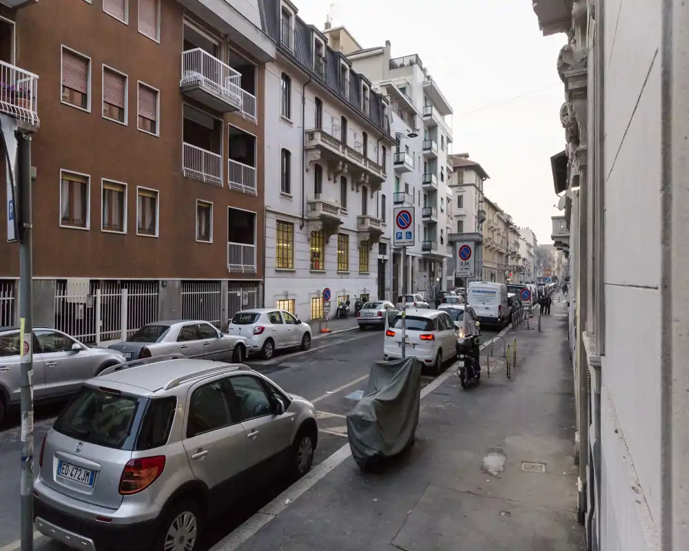 Vista a nivel de calle de una vía urbana europea con coches aparcados, motocicletas y edificios residenciales de altura media a ambos lados; luz vespertina suave y acera visible.