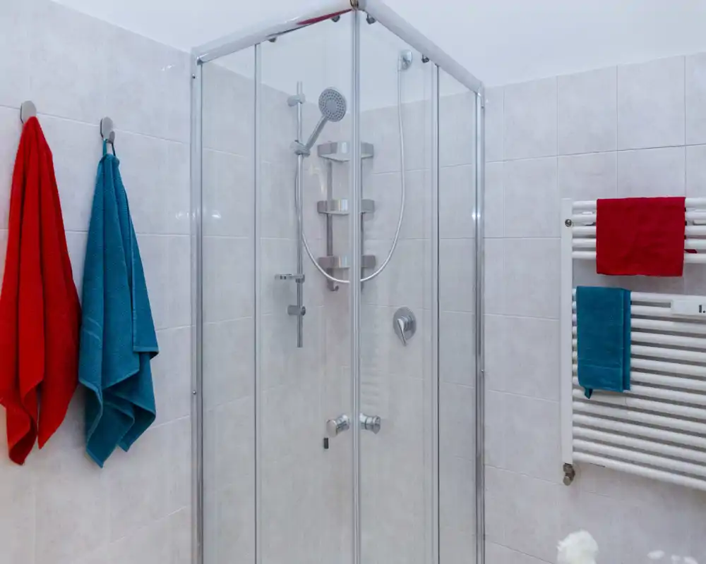Baño moderno y limpio con ducha de esquina de cristal, grifería cromada y paredes alicatadas, realzado por toallas de colores colgadas y un toallero calefactado.
