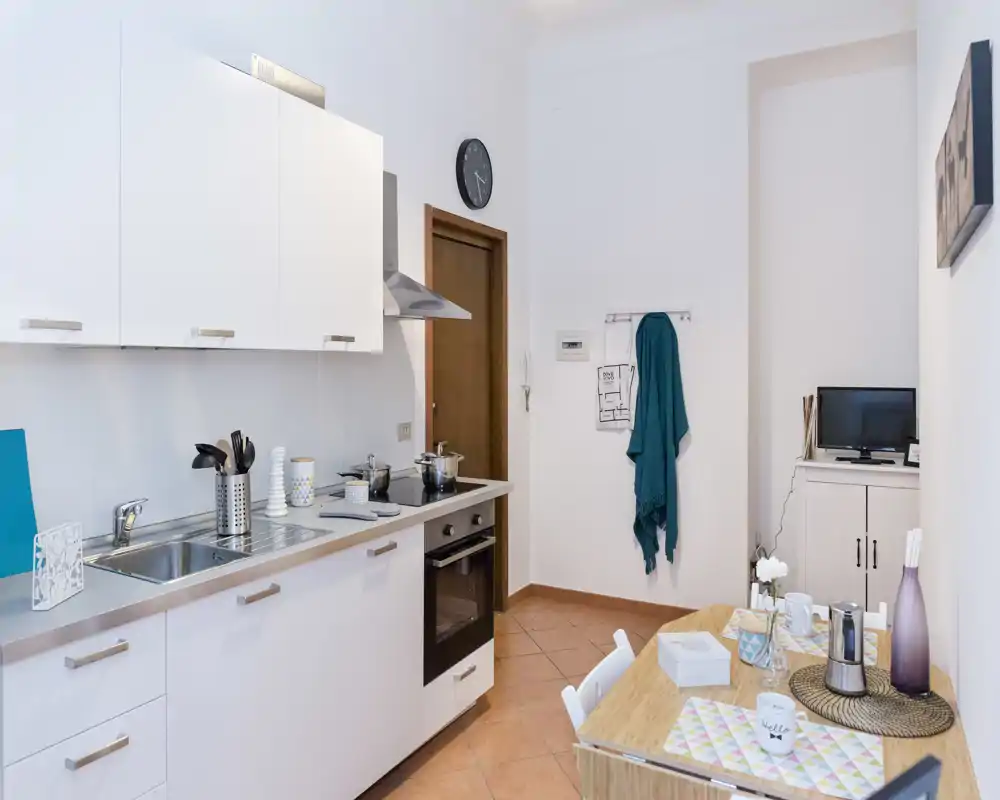 Cocina compacta y luminosa con muebles blancos, fregadero de acero, cocina y una pequeña mesa preparada para dos — ordenada y bien presentada para alquileres vacacionales.