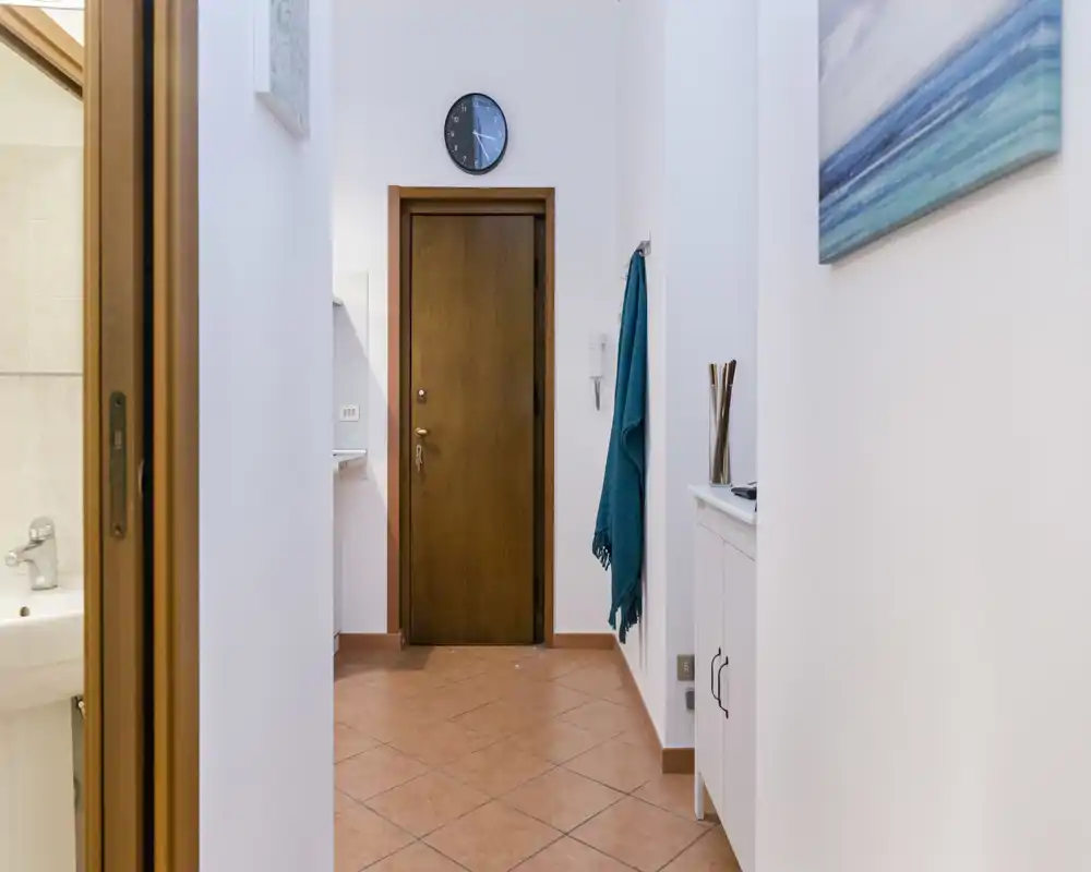 Entrada de apartamento luminosa y ordenada con suelo de baldosa, puerta de madera, reloj de pared y pequeño mueble — perfecta primera impresión.
