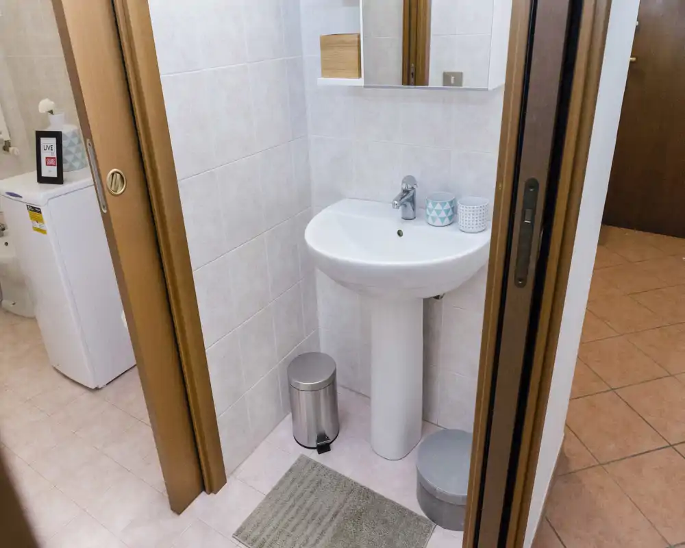 Pequeño baño limpio con lavabo de pedestal, espejo y azulejos rosa claro. La imagen muestra el área del lavabo ordenada y se aprecia el inodoro en la zona contigua.