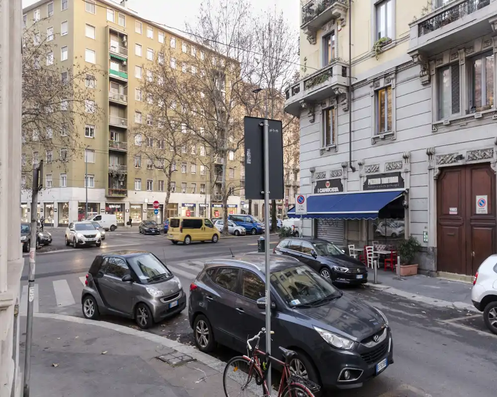 Escena urbana a nivel de calle con coches aparcados, una pequeña cafetería con toldo azul y edificios residenciales alrededor; fotografía diurna clara adecuada para mostrar la ubicación y el entorno.