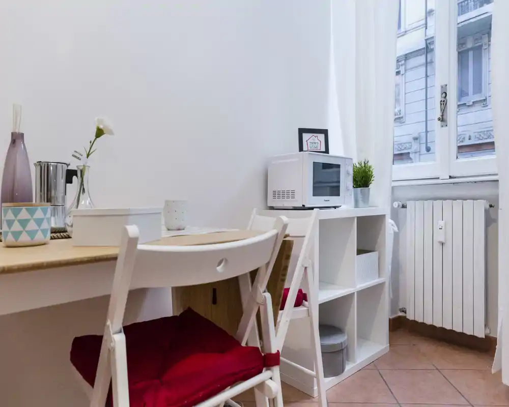 Pequeño rincón de cocina luminoso con mesa, sillas plegables, microondas y estantería — compacto y ordenado, ideal para estancias cortas.