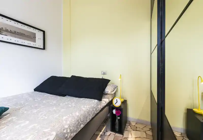 Compact, well-lit bedroom with a double bed, patterned duvet, black pillows and a mirrored wardrobe creating depth. A small bedside table with a yellow lamp and clock adds a cheerful accent.