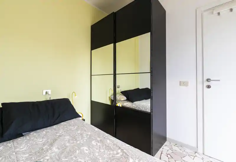 Compact, modern bedroom with a patterned duvet, black pillows and a mirrored wardrobe that visually expands the space. The room is tidy, well lit and furnished for comfortable short stays.