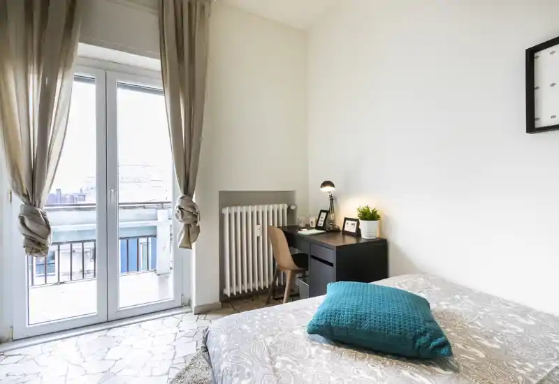 Bright, cozy bedroom with a comfortable bed, textured cushion and a small workspace by the window leading to a balcony. Soft natural light and neutral decor make this room inviting for restful stays.