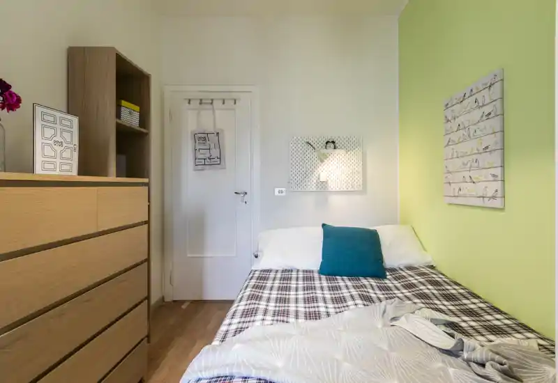 Dormitorio compacto y bien decorado con cama doble junto a una pared verde claro, cómoda de madera y luz ambiental suave — luminoso y acogedor para los huéspedes.