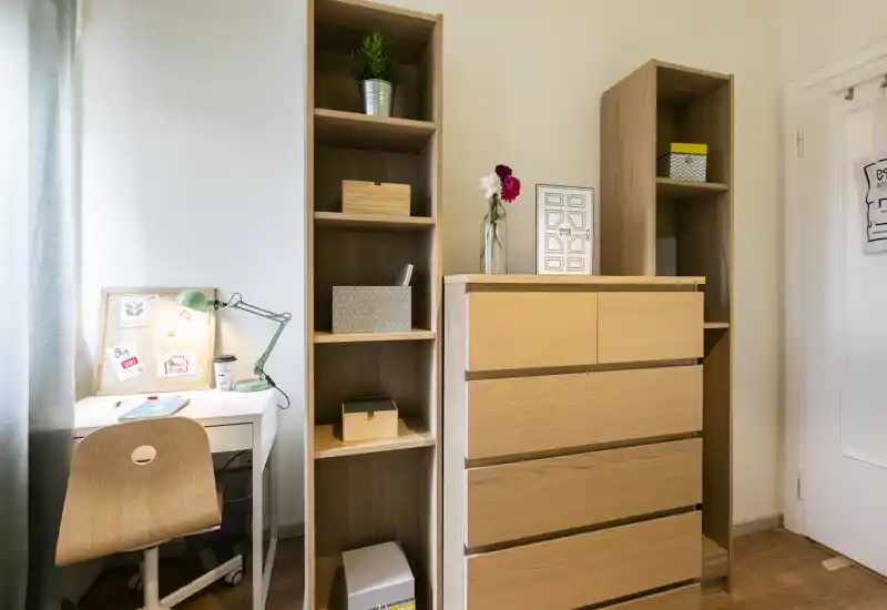 Rincón de dormitorio ordenado con cómoda de madera, estanterías abiertas y pequeño escritorio con lámpara: acabados cálidos y estilo cuidado que crean un espacio acogedor y funcional.