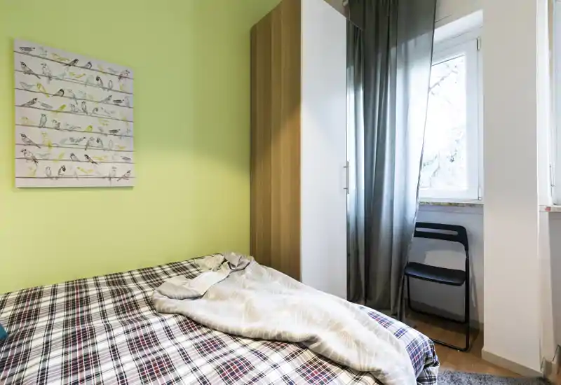 Dormitorio compacto y luminoso con cama doble con ropa de cama estampada, un armario alto y ventana con cortina fina que crean un ambiente acogedor y aireado.