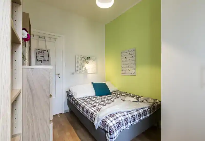 Dormitorio pequeño y bien decorado con cama doble frente a una pared verde, textiles acogedores y una lámpara que crea un ambiente agradable.