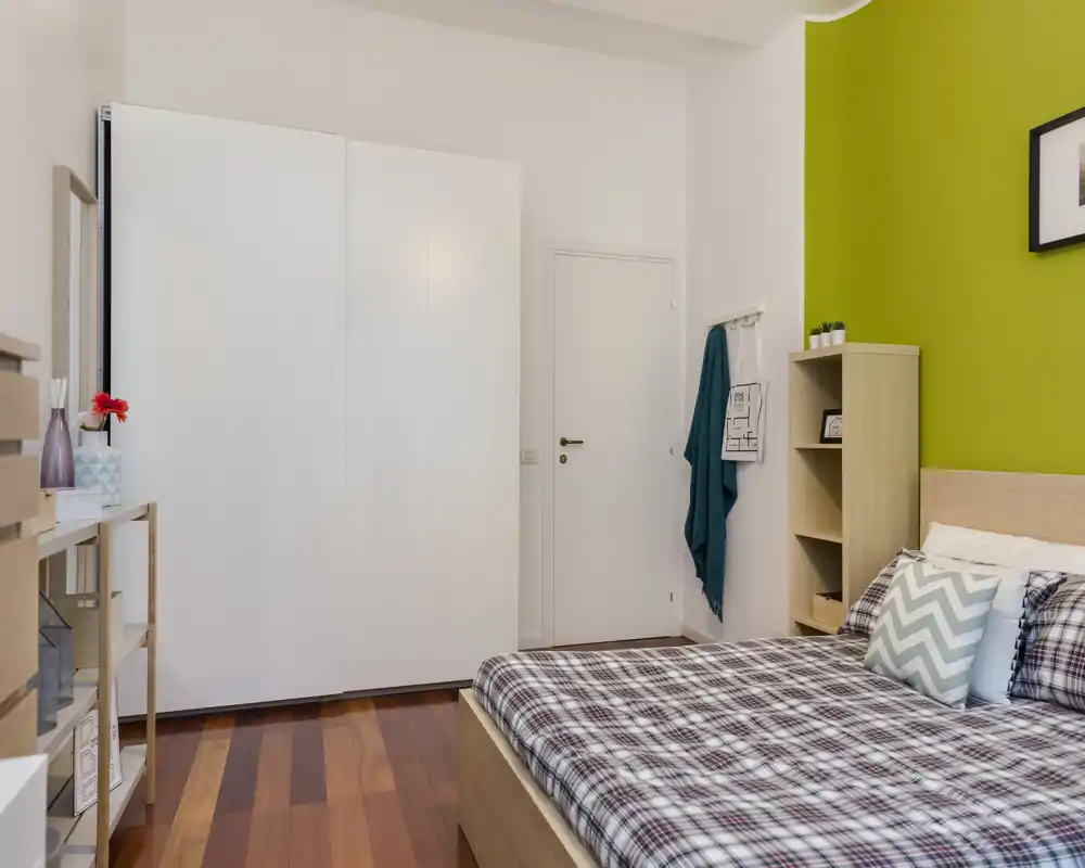 Camera da letto moderna e ben illuminata con letto matrimoniale confortevole, biancheria a fantasia e una parete verde brillante. Armadio a muro e scaffalature offrono spazio pratico; il pavimento in legno caldo aggiunge comfort.
