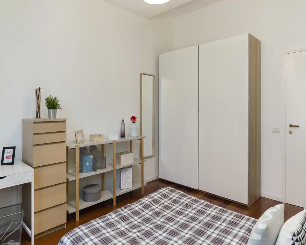 Camera da letto luminosa e moderna con letto ben rifatto in primo piano, mobili in legno chiaro e un grande armadio bianco per un aspetto pulito e minimal.