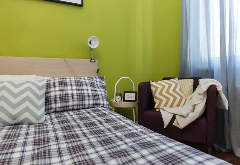 Camera da letto luminosa e moderna con biancheria a fantasia, poltrona d'accento e comodino; luce naturale piacevole e parete verde brillante.