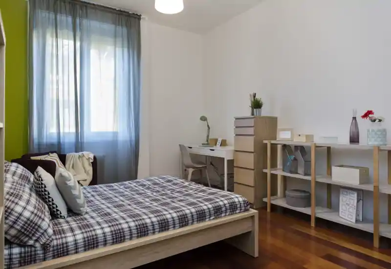 Camera da letto luminosa e ben arredata con letto in legno, biancheria a quadri, scrivania e scaffali—ideale per una persona o ospiti.