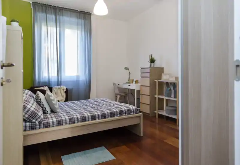 Camera da letto luminosa e ordinata con letto in legno, copriletto a quadri, scrivania e contenitori: perfetta come stanza singola o per gli ospiti.