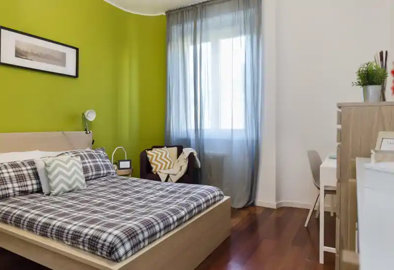 Camera da letto luminosa e di stile con letto in legno, biancheria a fantasia e luce naturale soffusa da una grande finestra. La parete verde e i tessili accoglienti creano un'atmosfera invitante perfetta per gli affitti.