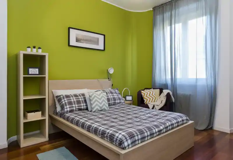 Camera da letto luminosa e ben allestita con un letto matrimoniale contro una parete verde, biancheria a fantasia e una poltrona accogliente vicino alla finestra.